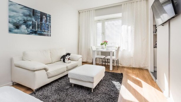 A modern living room with a white sofa, a black and white dog, a dining table with chairs, a TV mounted on the wall, and a window with white curtains. - Go Happy Home Apt Vuorimiehenkatu 14 (Helsinki)