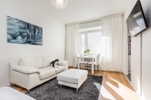 A modern living room with a white sofa, a black and white dog, a dining table with chairs, a TV mounted on the wall, and a window with white curtains. - Go Happy Home Apt Vuorimiehenkatu 14 (Helsinki)