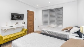 A modern bedroom with a bed, a sofa, a TV, and a window with shutters. - Goldbeaters House (Edgware)