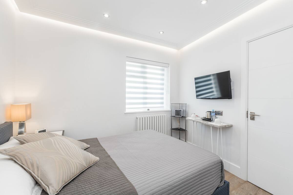 A modern bedroom with a bed, a TV mounted on the wall, a bedside table with a lamp, and a window with blinds.