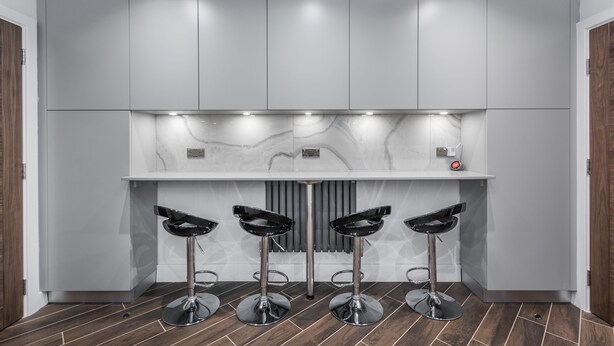 A modern kitchen with grey cabinets, a marble backsplash, and three black bar stools. - Goldbeaters House (Edgware)