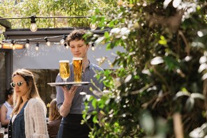 A person carrying a tray with two glasses of beer and a bottle, outdoors with string lights and greenery in the background. - Aragon House (London)