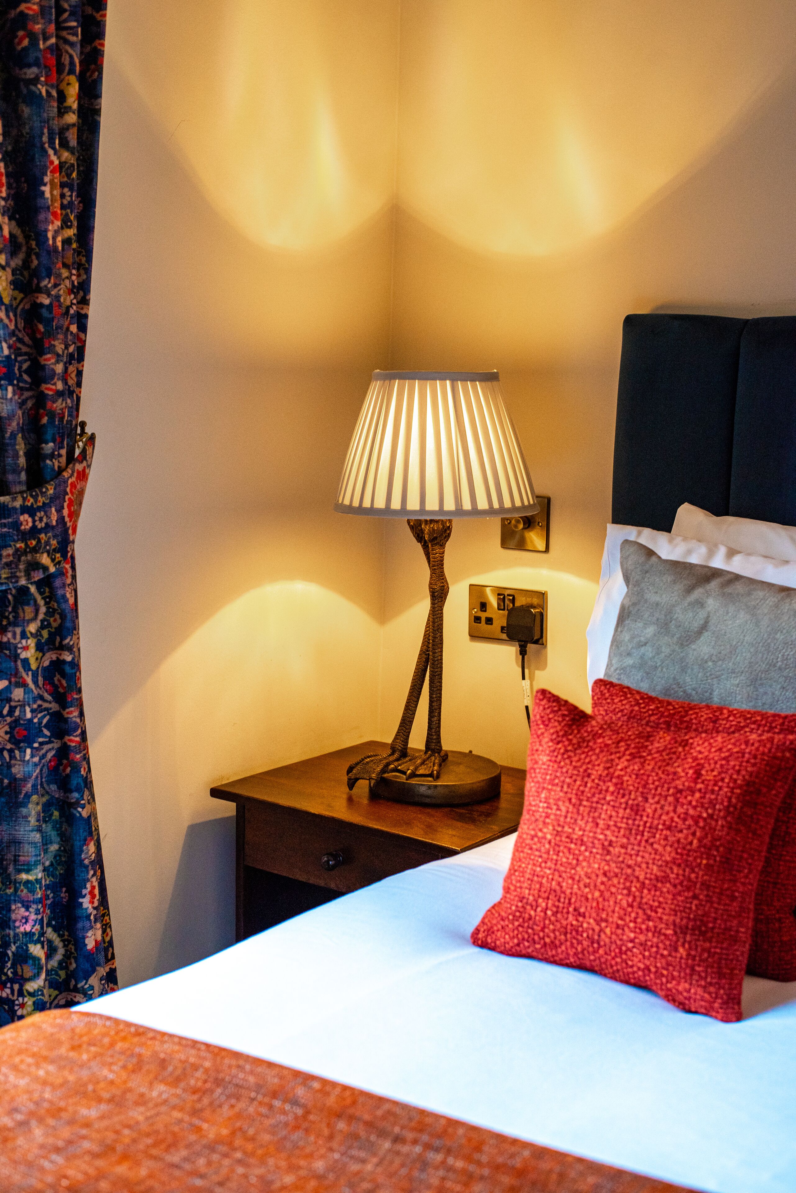 Een net opgemaakt bed met een rood en grijs kussen, een lamp op een nachtkastje en een stopcontact aan de muur.