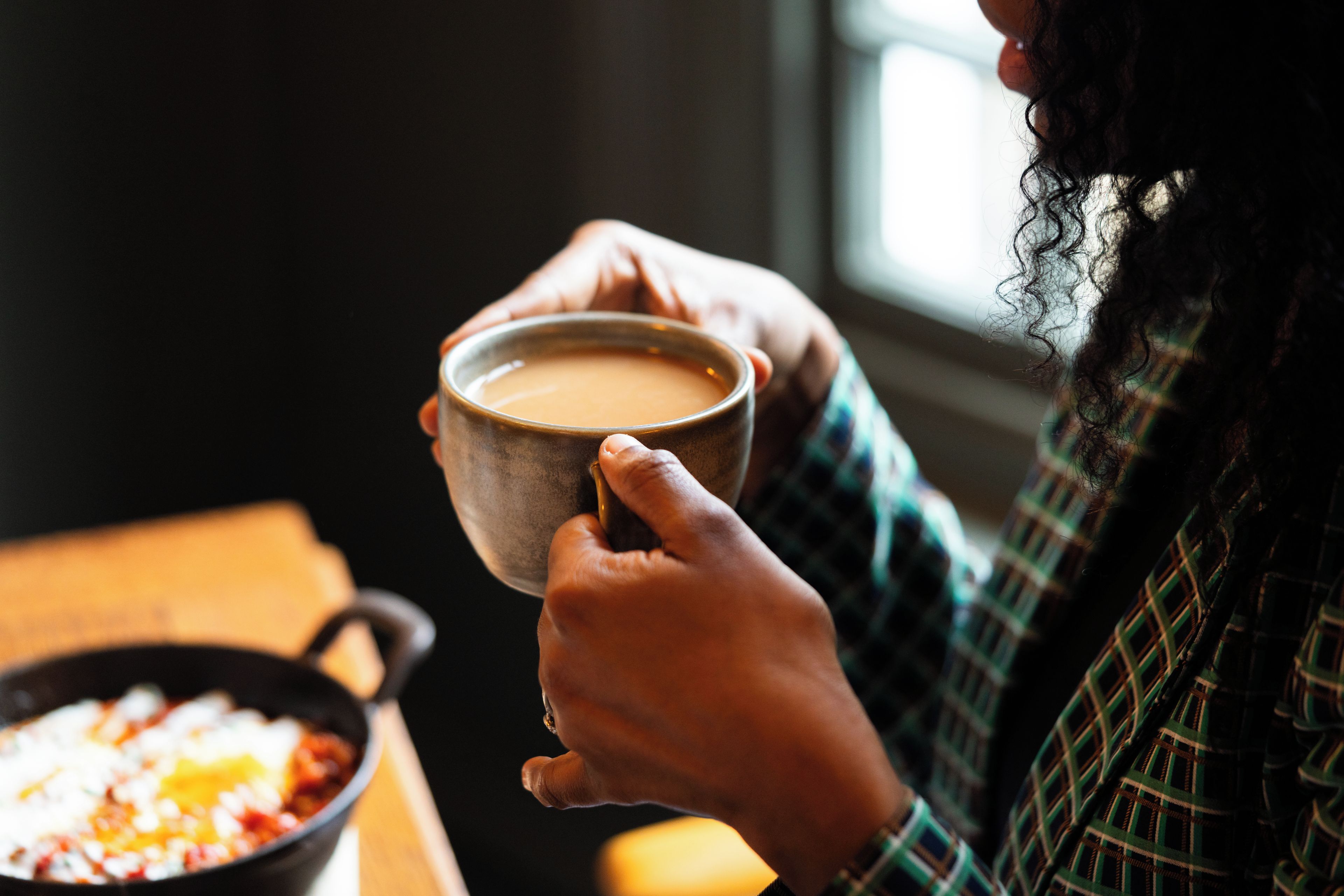 A person holding a mug, with a pan of food on a table in the background.