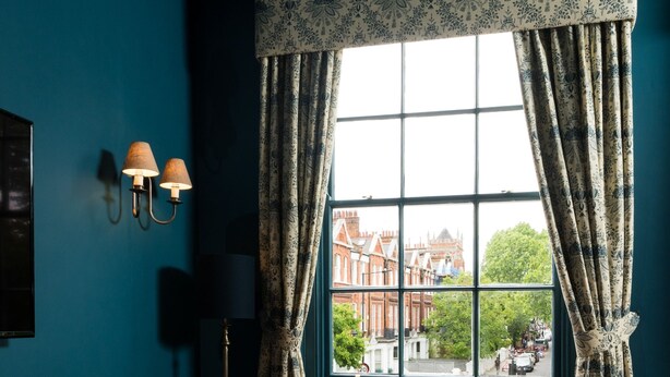 A room with a teal wall, a brown sofa, a white ottoman, and a window with patterned curtains. - Aragon House (London)