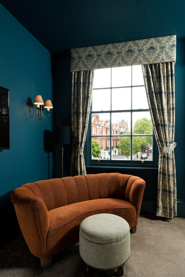 A room with a teal wall, a brown sofa, a white ottoman, and a window with patterned curtains. - Aragon House (London)