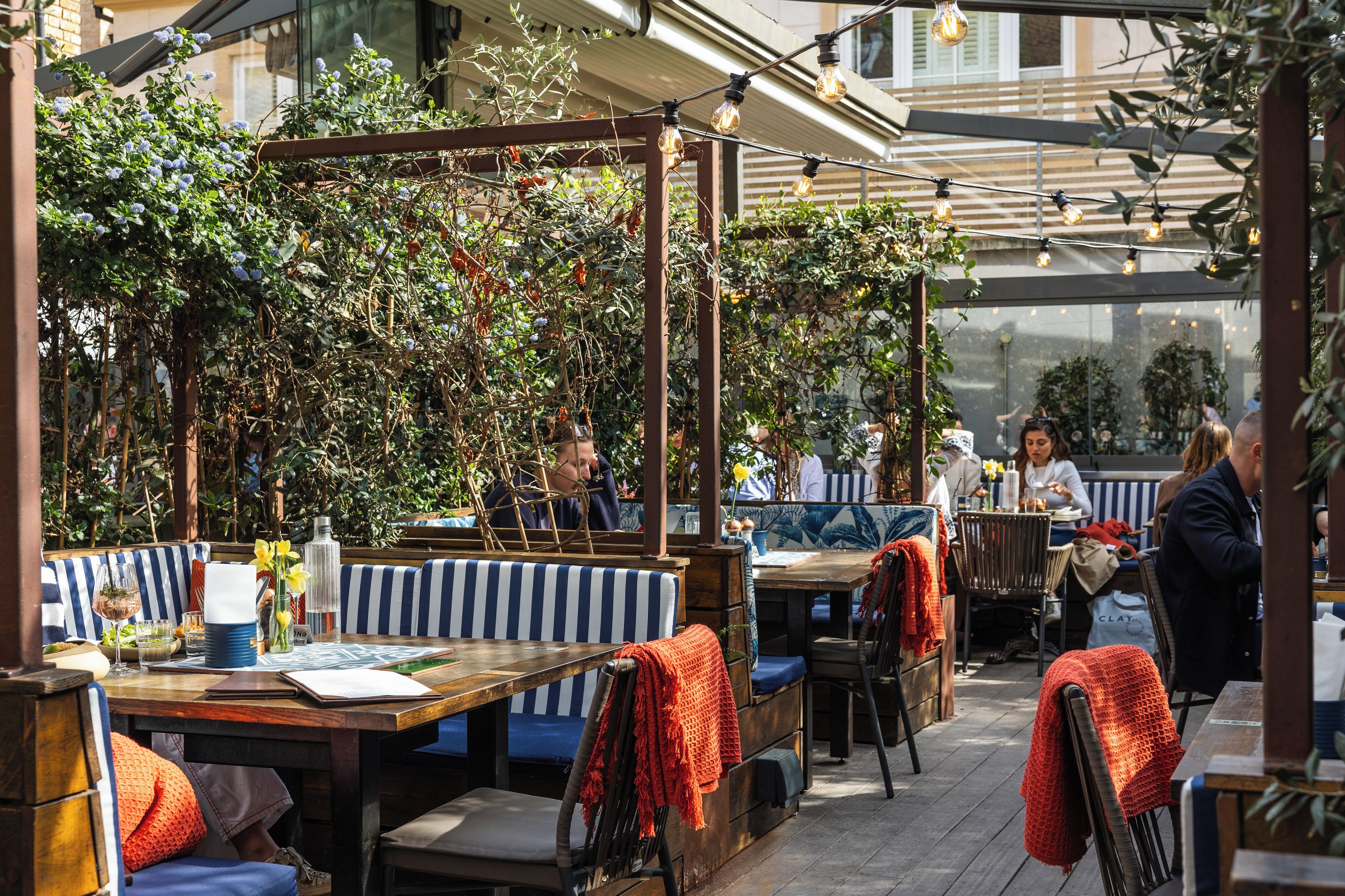 An outdoor seating area with tables and chairs, some with red blankets, and a wooden pergola with climbing plants.