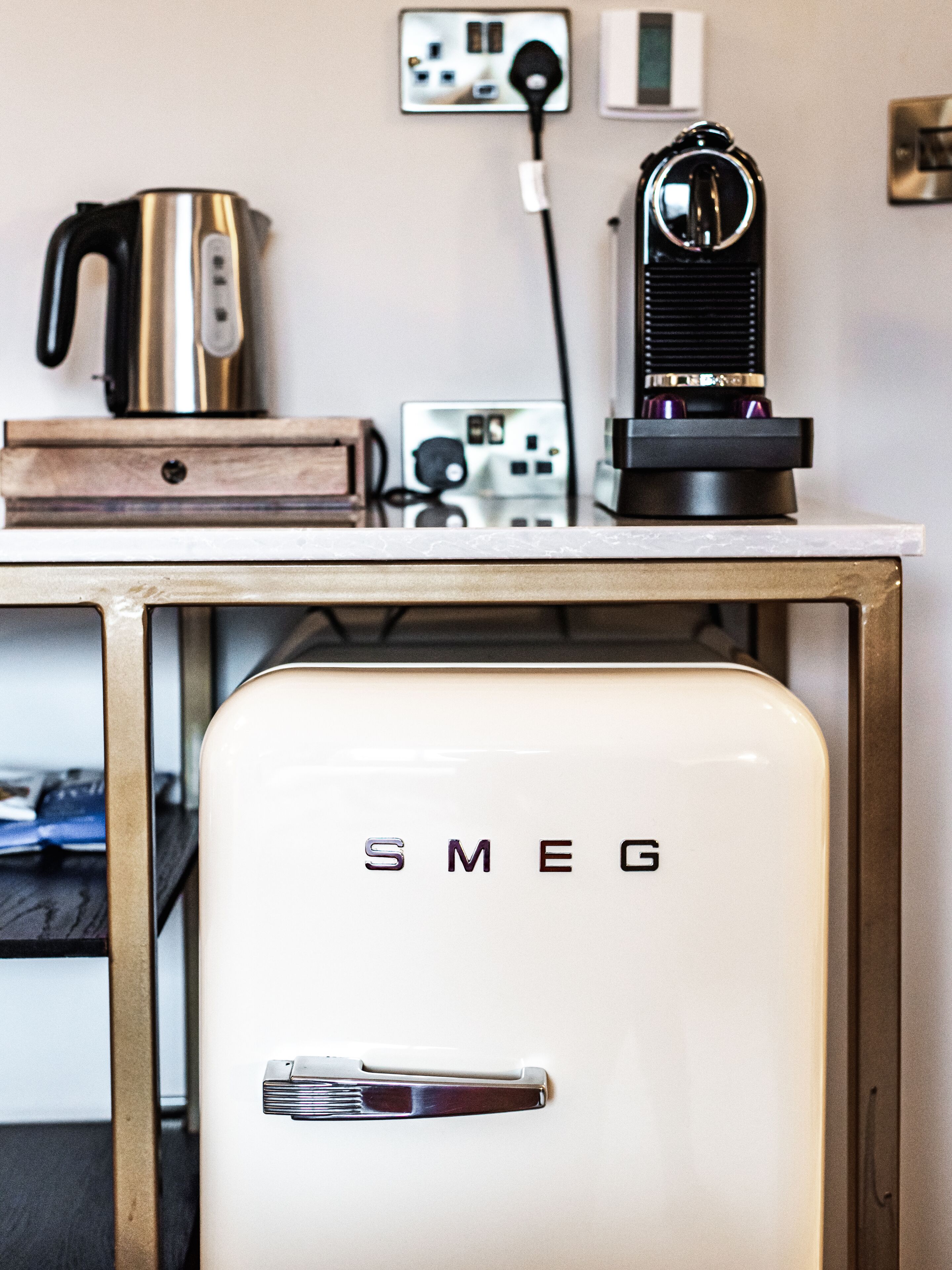 A kitchenette with a white Smeg refrigerator, a coffee maker, and a kettle on a wooden table.