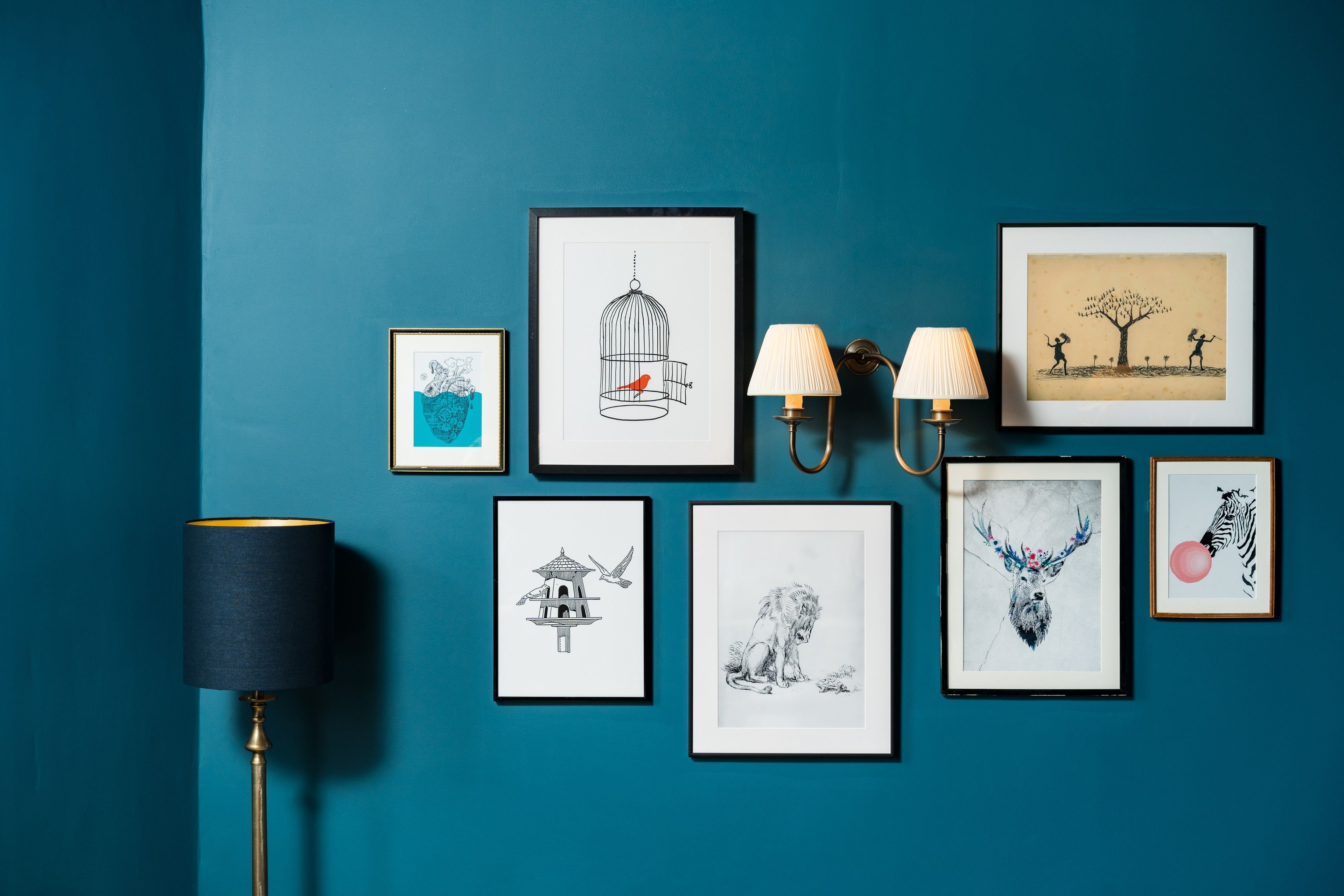 A teal wall with eight framed artworks, a floor lamp, and a wall-mounted lamp.