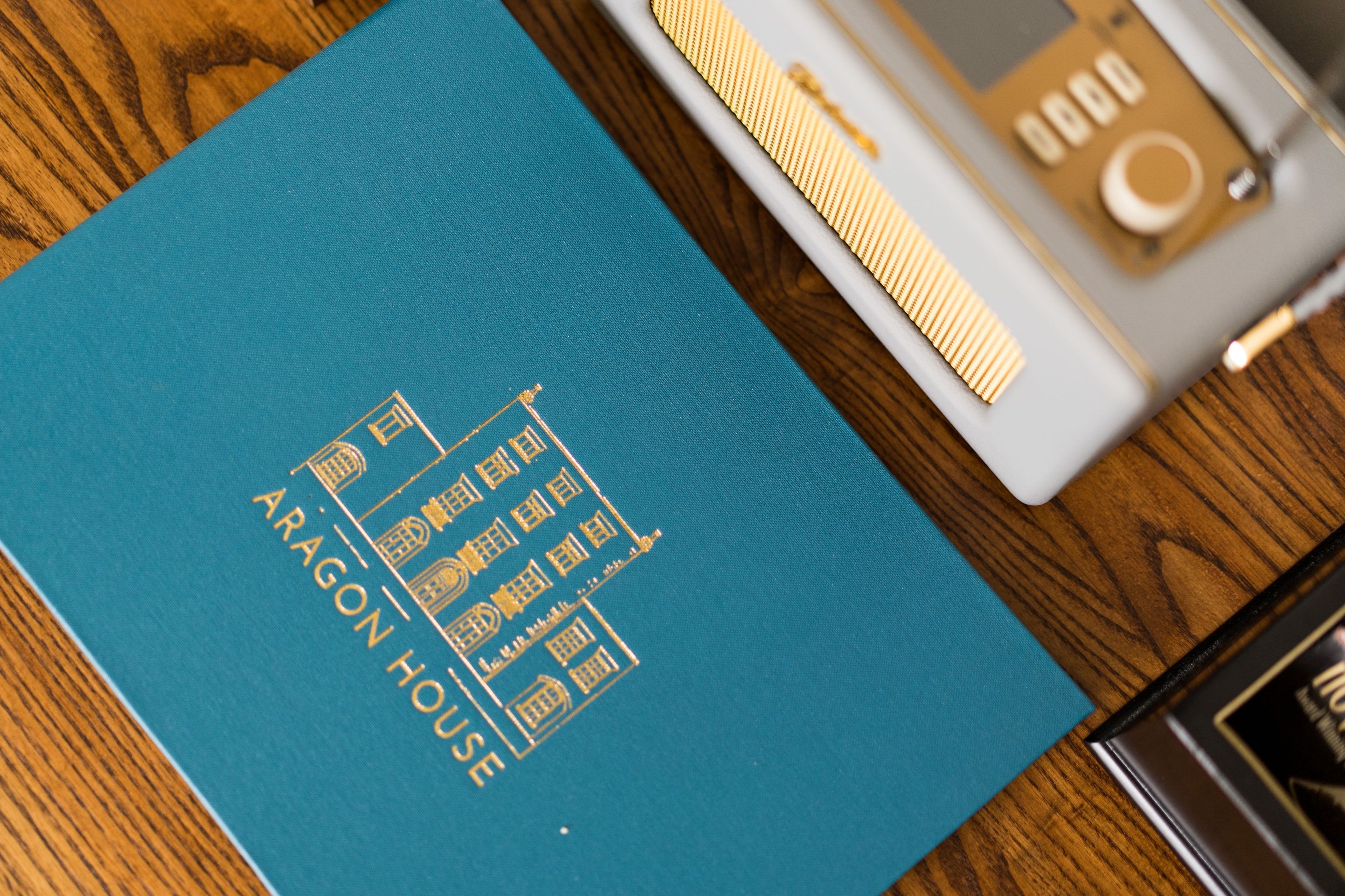A blue book titled "ARAGON HOUSE" with gold embossed text and symbols, placed on a wooden surface next to a white and gold object.