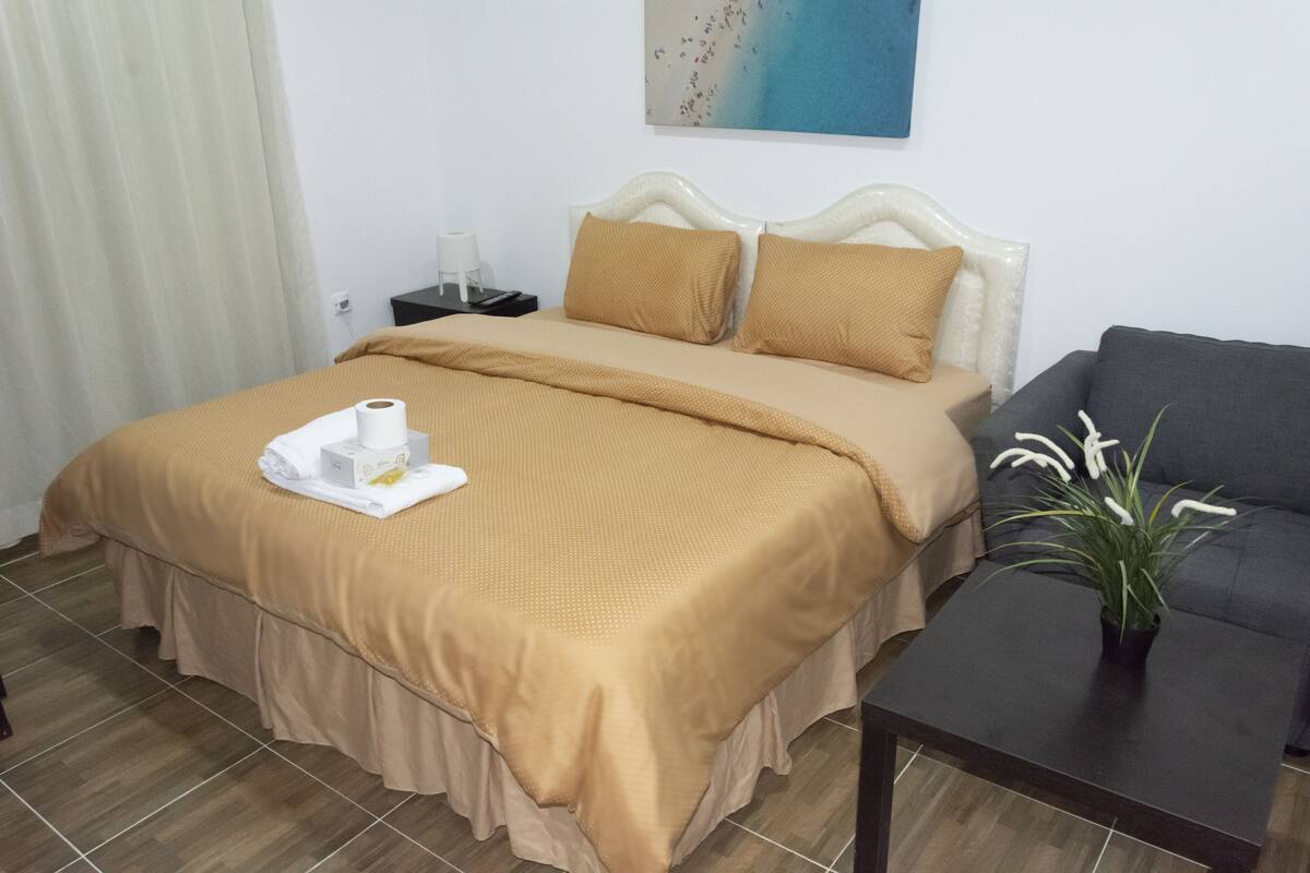 A hotel room with a bed, a bedside table, a small table with a plant, and a painting on the wall.