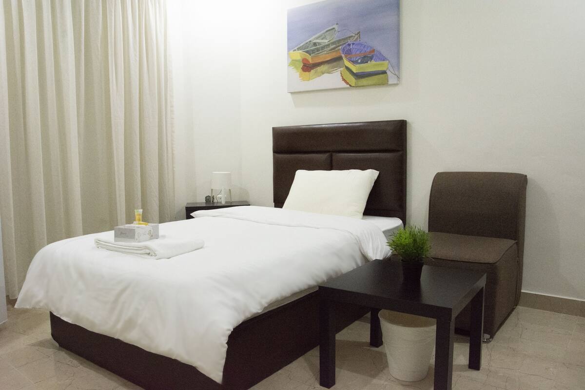 A neatly made bed with a white comforter and pillows, a small bedside table with a lamp, a chair, and a painting of boats on the wall.