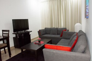 Una sala de estar con televisor, un sofá gris, un cojín rojo, una silla de madera y una mesita con un control remoto.
