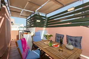 A wooden table set with plates, a bottle, and glasses, surrounded by blue chairs and cushions, under a shaded area with a view of buildings. - Chania Escapes Old Town-Casa Barchetta- Adults Only (Chania)