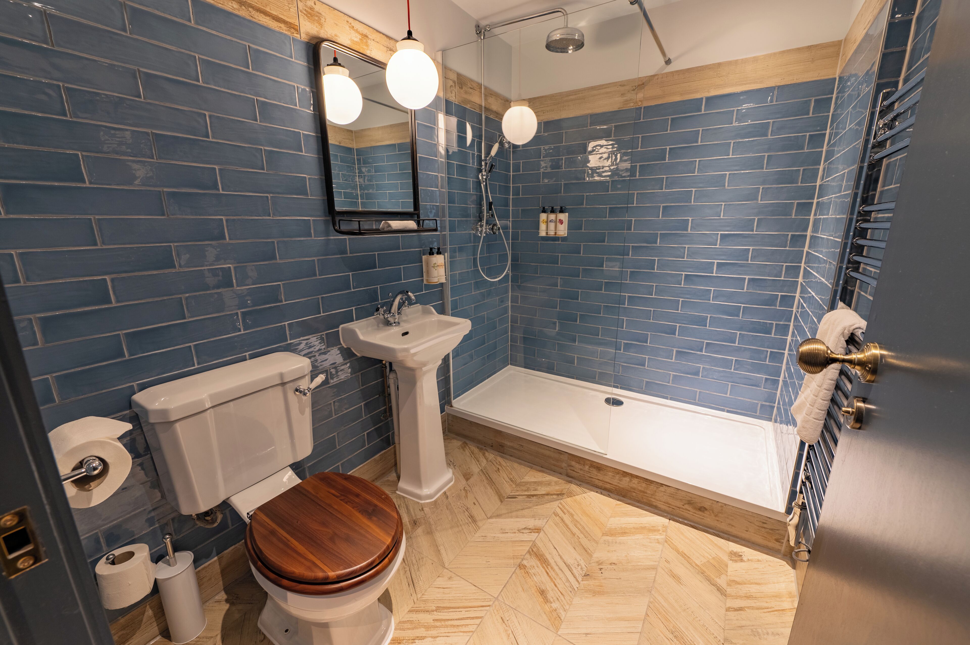 A bathroom with blue tiled walls, a wooden floor, a white sink, a toilet, and a shower.