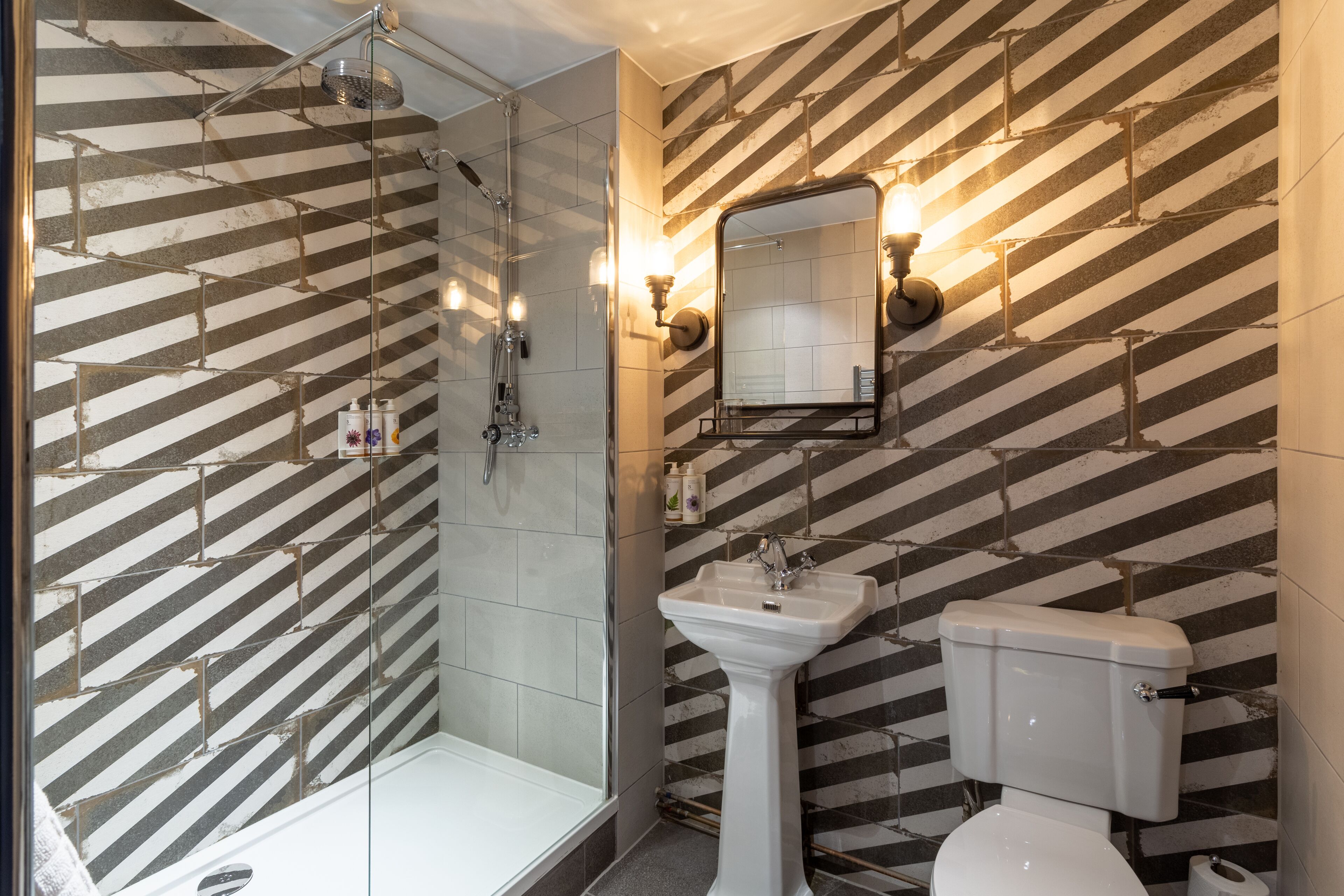 A bathroom with a shower, sink, and toilet, featuring a striped tile backsplash and a mirror with wall-mounted lights.