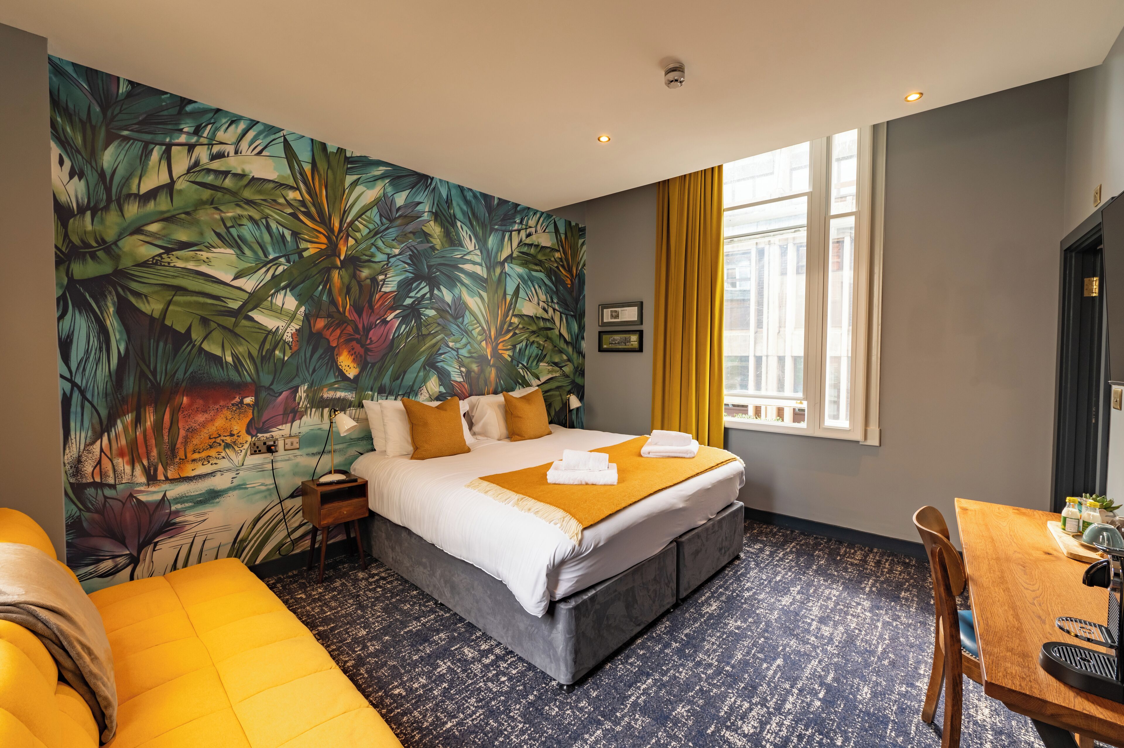 Una habitación de hotel con cama, sofá amarillo, mesa de comedor y un gran mural tropical en la pared.