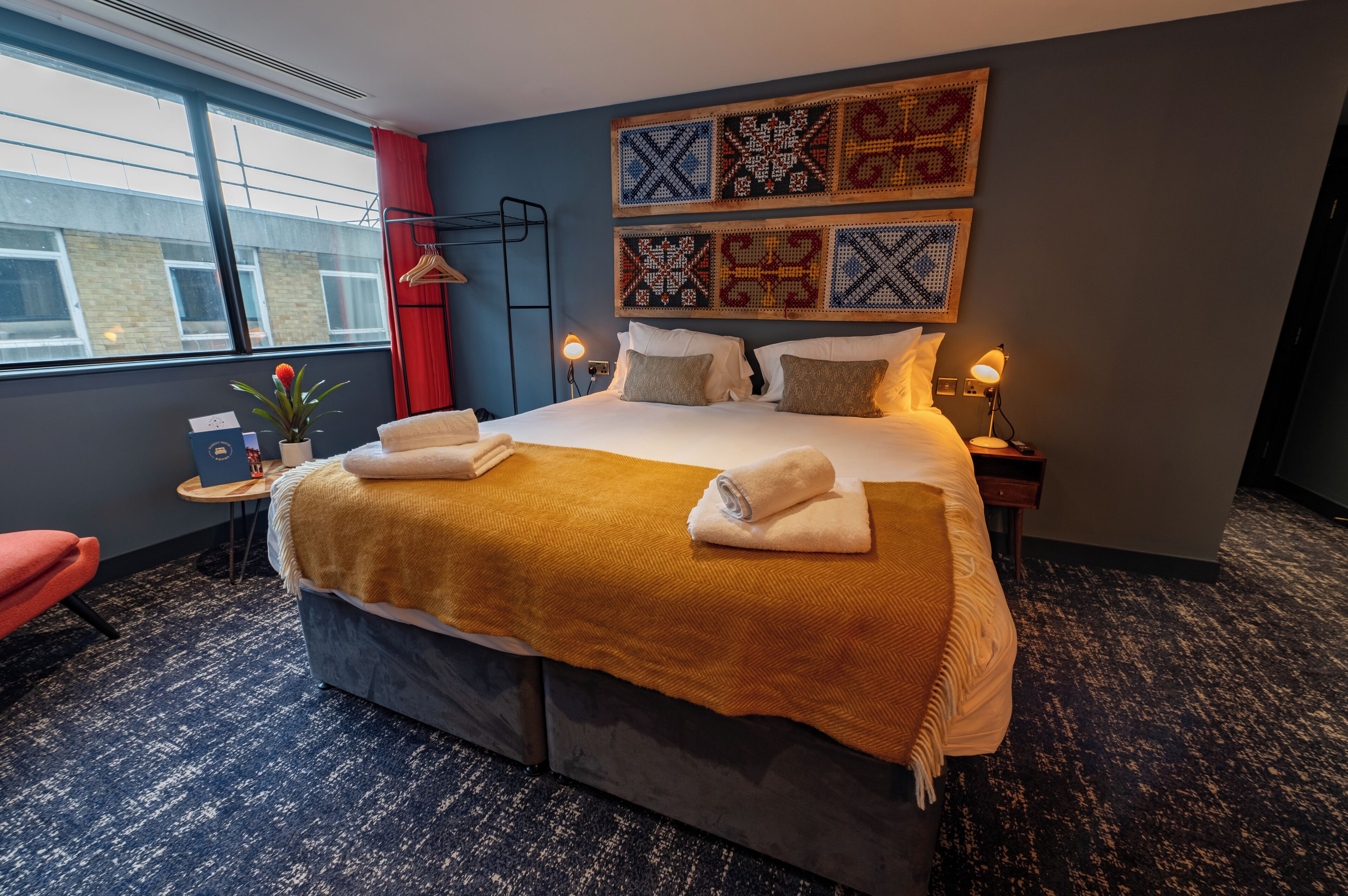 A hotel room with a large bed, a bedside table with a lamp, a wall-mounted artwork, and a window with a view of buildings.