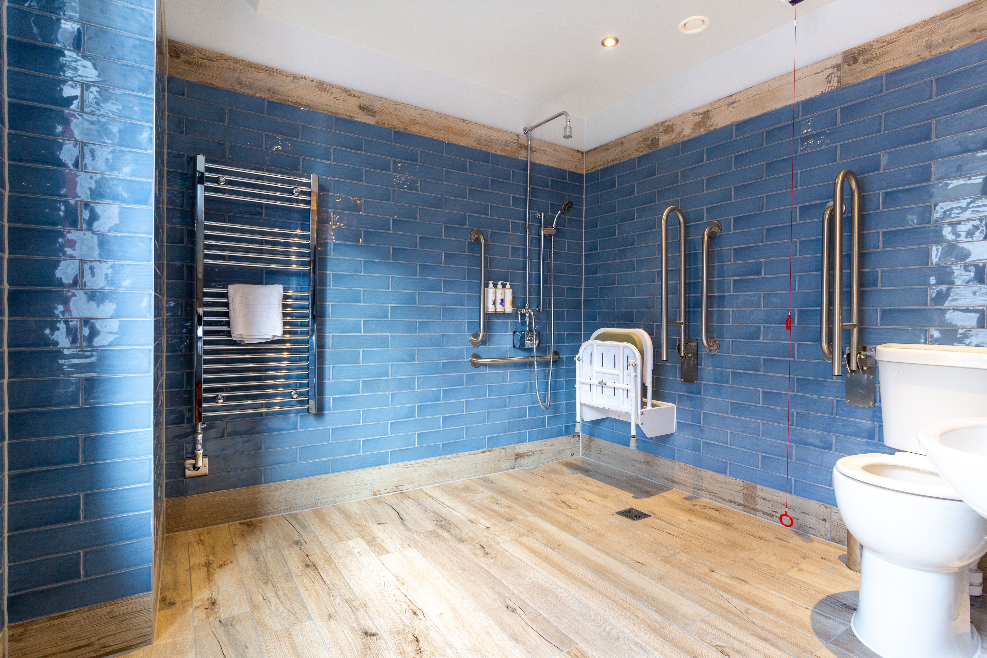 Un baño con paredes de azulejos azules, suelo de madera, inodoro blanco, ducha con cabezal de ducha manual y un perchero para toallas.