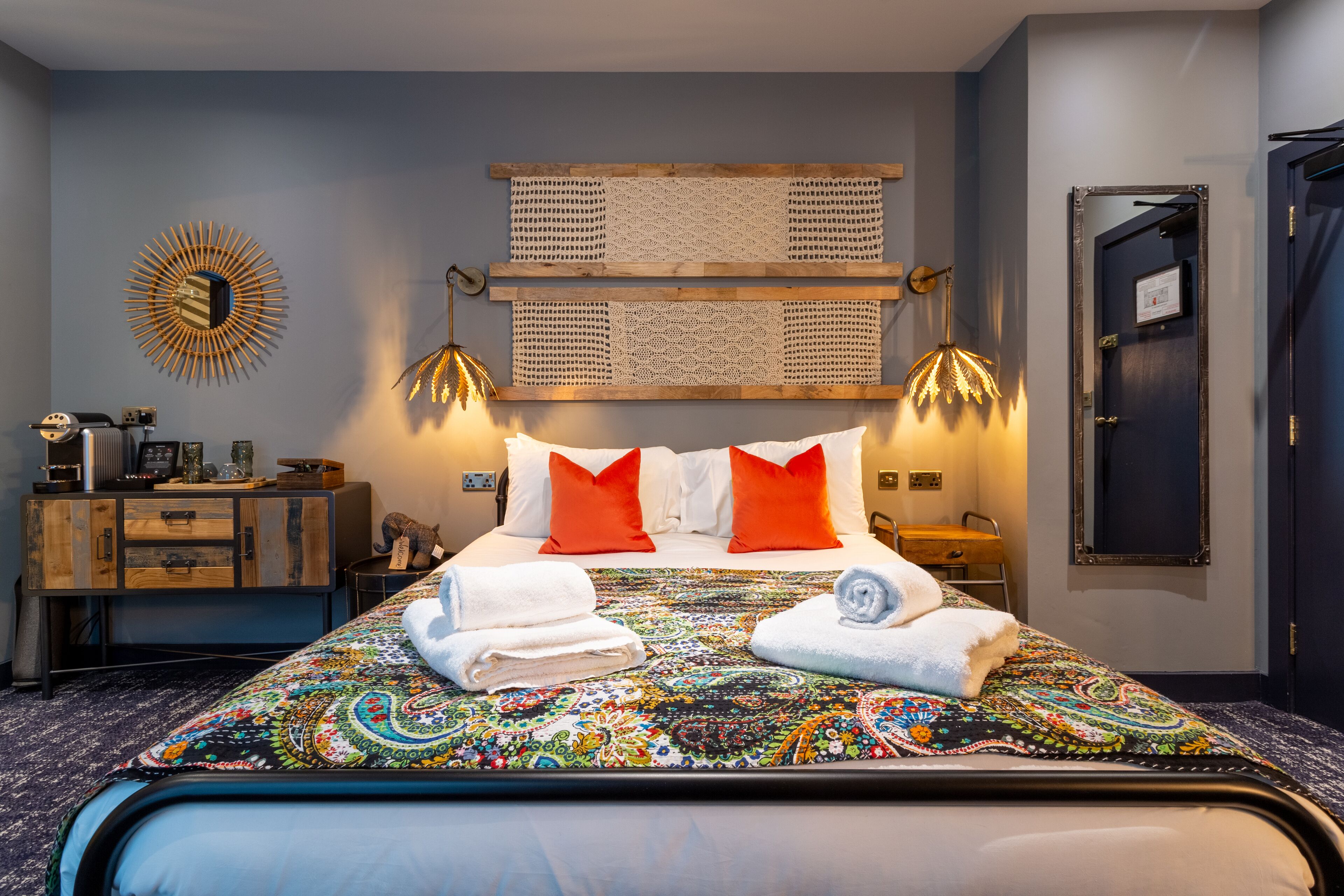 A neatly made bed with a colorful bedspread, two red and two white pillows, and two rolled towels. There is a wooden dresser with a mirror and a lamp on the left. The room has a blue wall with a decorative sunburst mirror and a woven wall hanging above the bed.