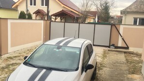 Un coche blanco aparcado en una entrada con valla, casas al fondo y cielo nublado.