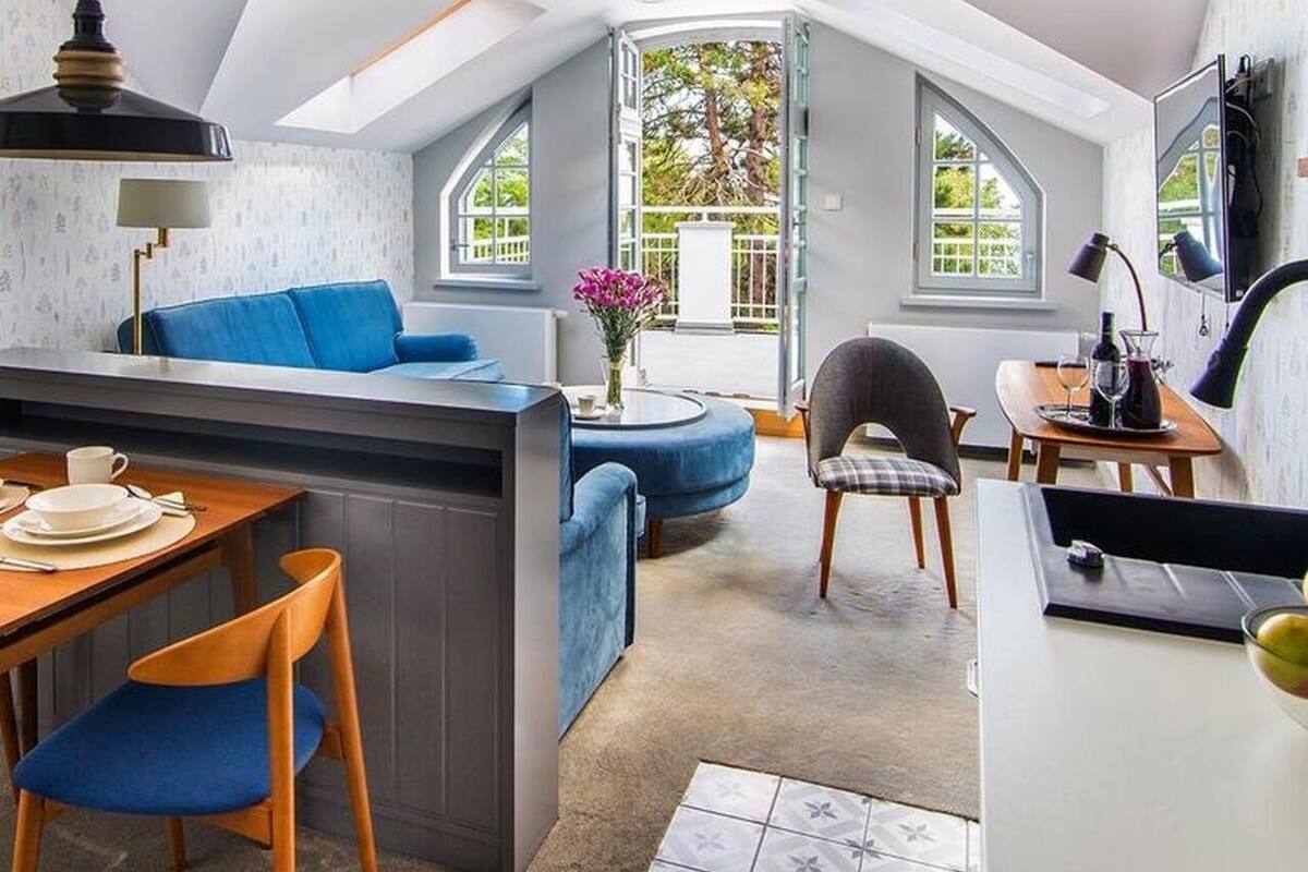 Un appartement moderne de style « loft », comprenant un coin repas, un canapé bleu, une petite cuisine et de grandes fenêtres offrant une vue sur l’extérieur.