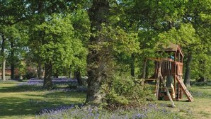 Een houten speeltuintje met een glijbaan en klimnet, omgeven door bomen en paarse bloemen.