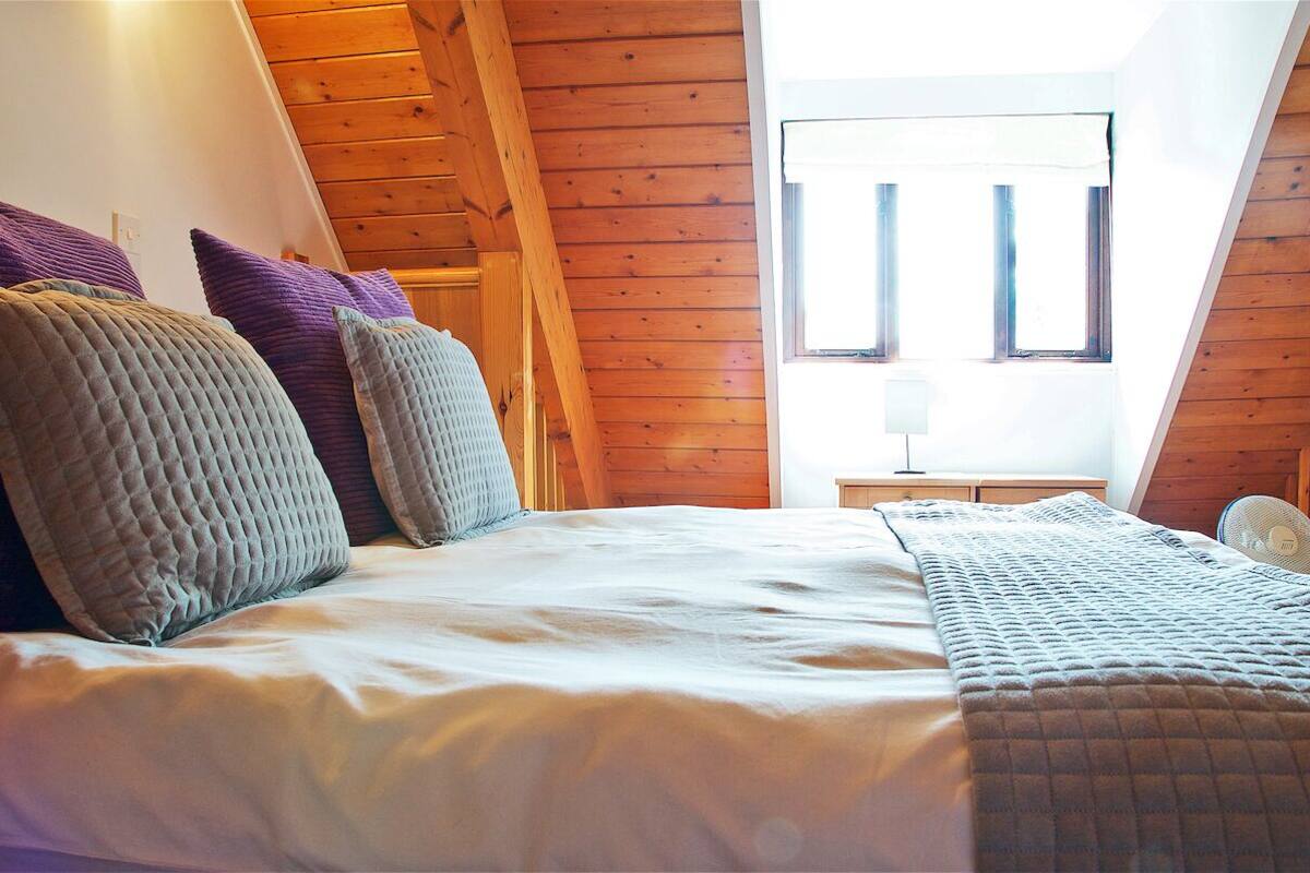 Een slaapkamer met een houten schuine daklijn, een bed met meerdere kussens, een nachtkastje met een lamp en een raam met zonwering.