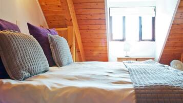 A bedroom with a wooden sloped ceiling, a bed with multiple pillows, a bedside table with a lamp, and a window with blinds.