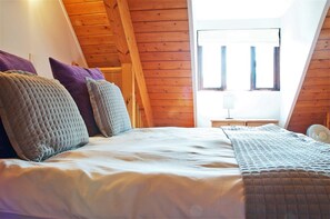 A bedroom with a wooden sloped ceiling, a bed with multiple pillows, a bedside table with a lamp, and a window with blinds. - Northlands Farm & Lakes (Chichester)