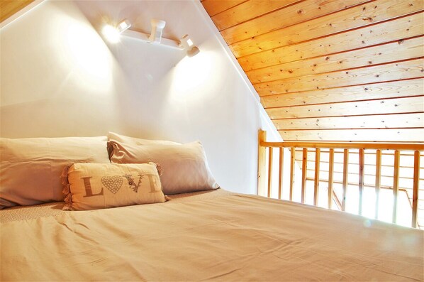 Un dormitorio con techo inclinado, vigas de madera y una cama con ropa de cama y almohadas color beige.