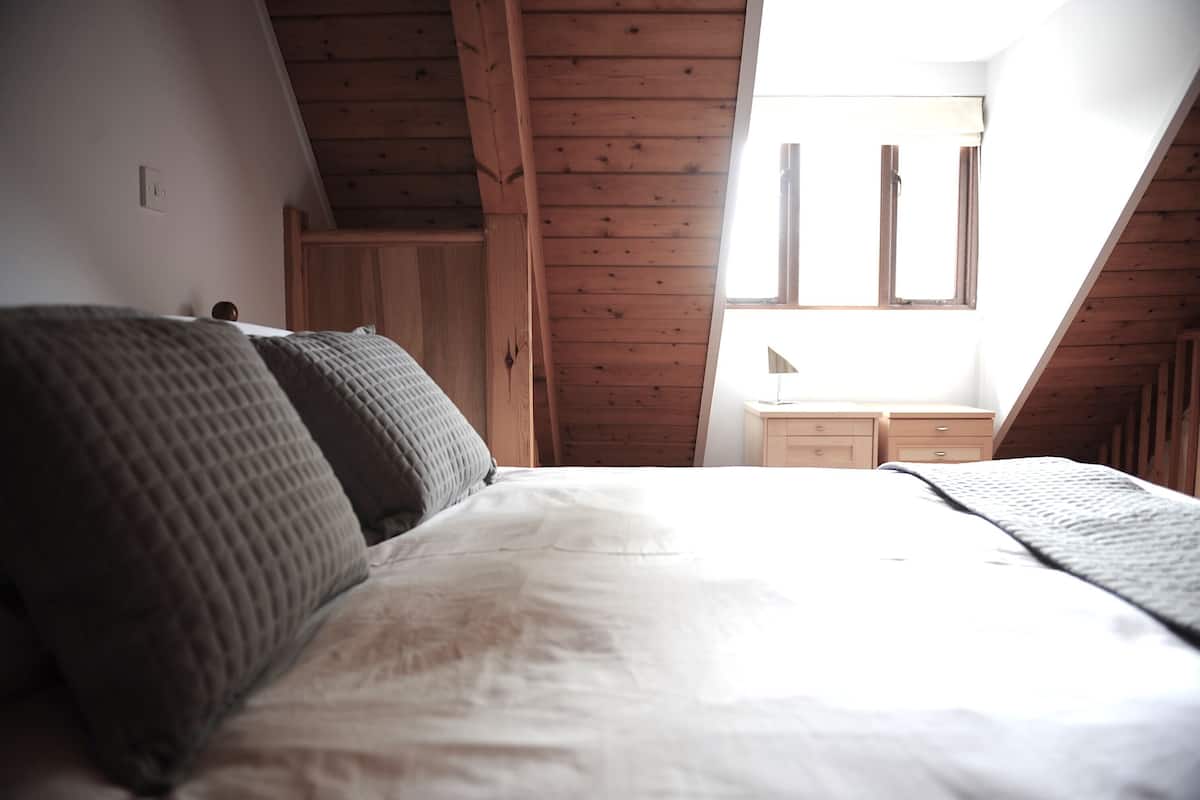Een slaapkamer met een schuine plafond, houten balken, een bed met een witte matras en twee kussens, een nachtkastje en een raam met uitzicht.