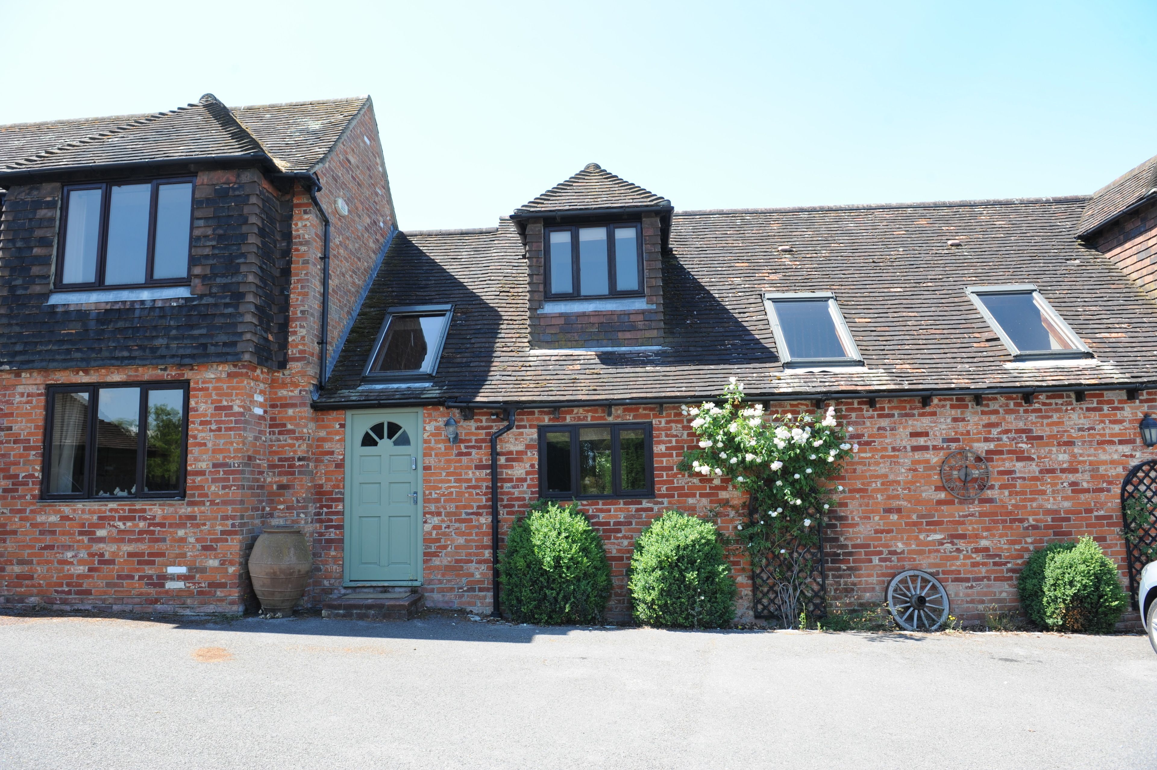 A two-story brick building with a green door, multiple windows, and a small garden area with a wheelbarrow and potted plants.