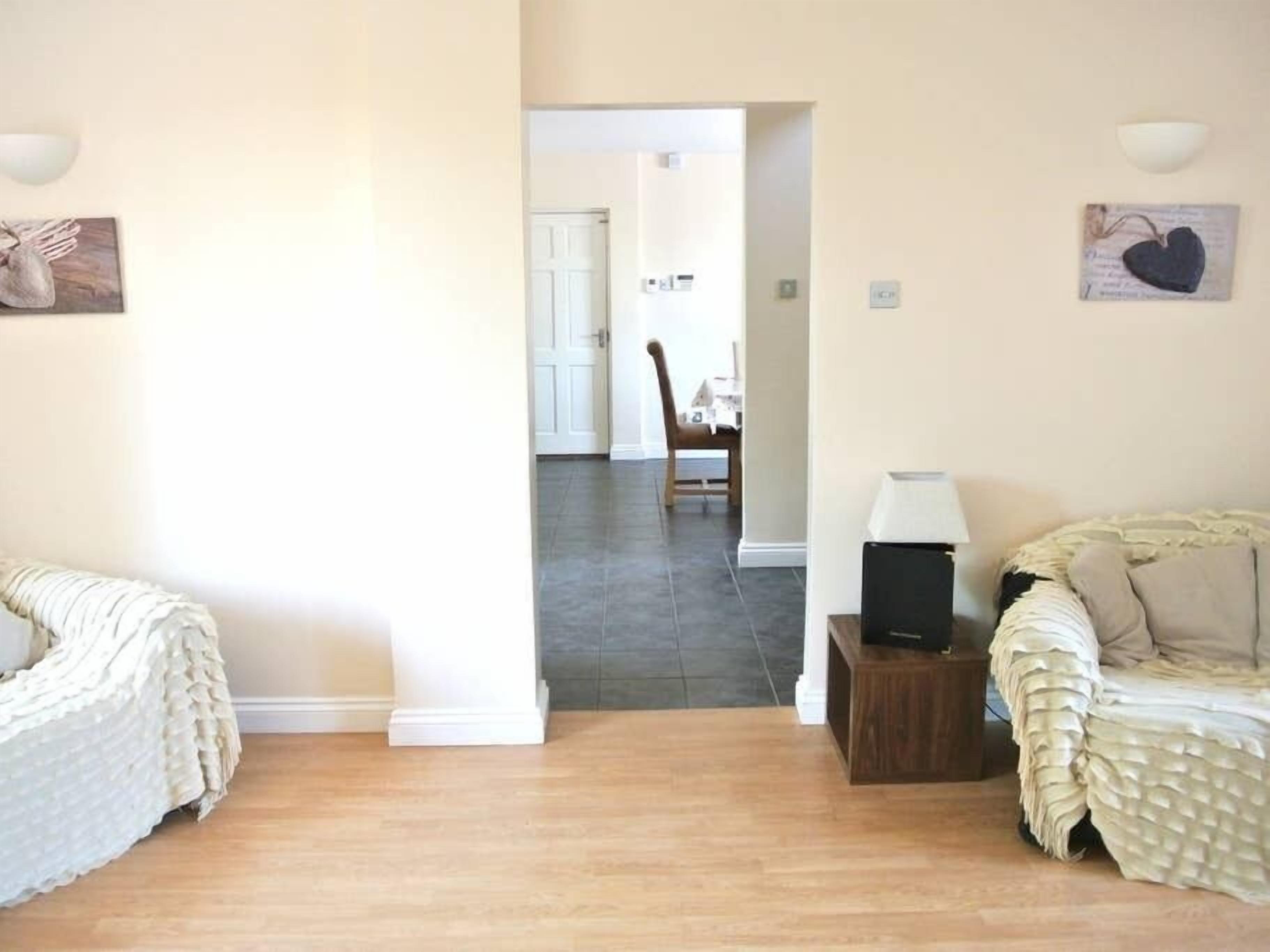 A room with a wooden floor, a bed with a textured white cover, a small wooden side table with a lamp, and a chair in the background.