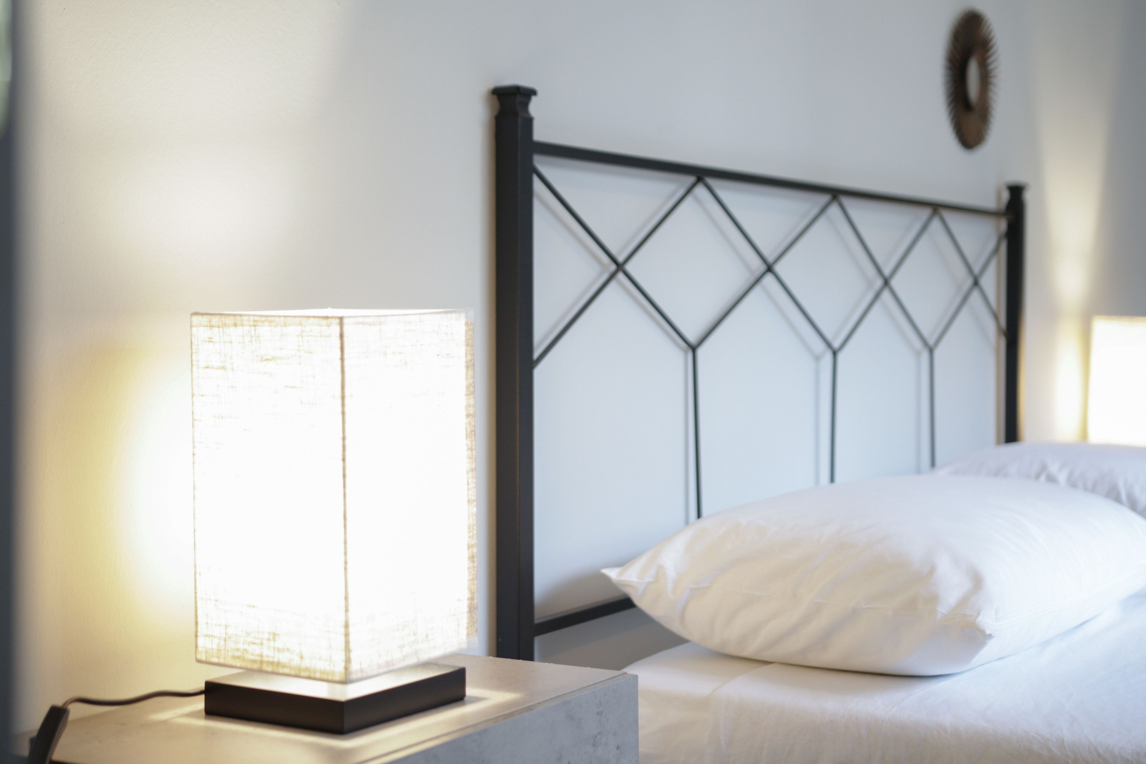 A neatly made bed with white linens and a single white pillow, a bedside table with a lamp, and a metal headboard.