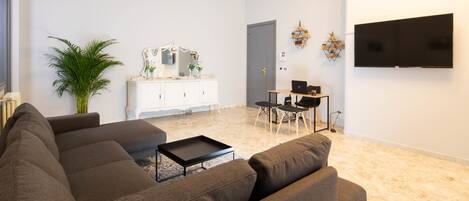 A modern living room with a sectional sofa, a small coffee table, a dining area with a table and chairs, a TV mounted on the wall, and a potted plant.