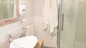 Un baño moderno con tocador de madera, un lavamanos blanco, cabina de ducha de vidrio y dos toallas blancas colgadas en la pared.