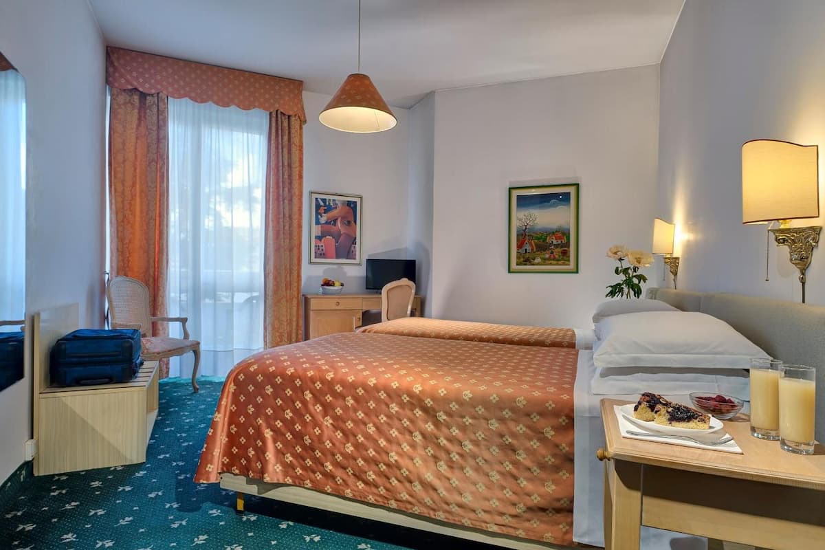 Una habitación de hotel con cama, escritorio, silla, maleta, mesita de noche con un plato de comida y bebidas, y ventana con cortinas.