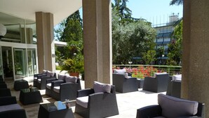 Een terras met rieten meubels en een glazen tafel, omgeven door groen en een uitzicht op gebouwen.