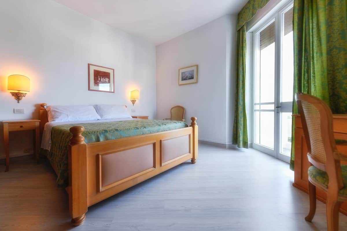 Un dormitorio con una cama de madera, dos lámparas de noche, una silla, una ventana con cortinas y un cuadro en la pared.