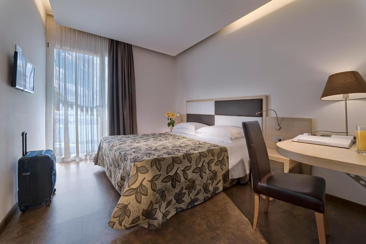 Una habitación de hotel con cama, escritorio, silla, lámpara, televisor y maleta.