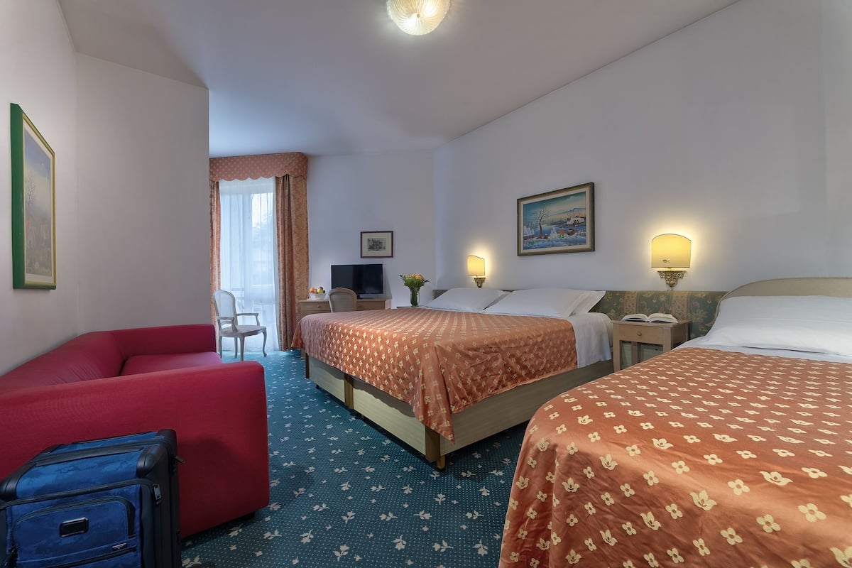 Habitación de hotel con dos camas, un sofá rojo, una maleta azul y un escritorio con una lámpara.