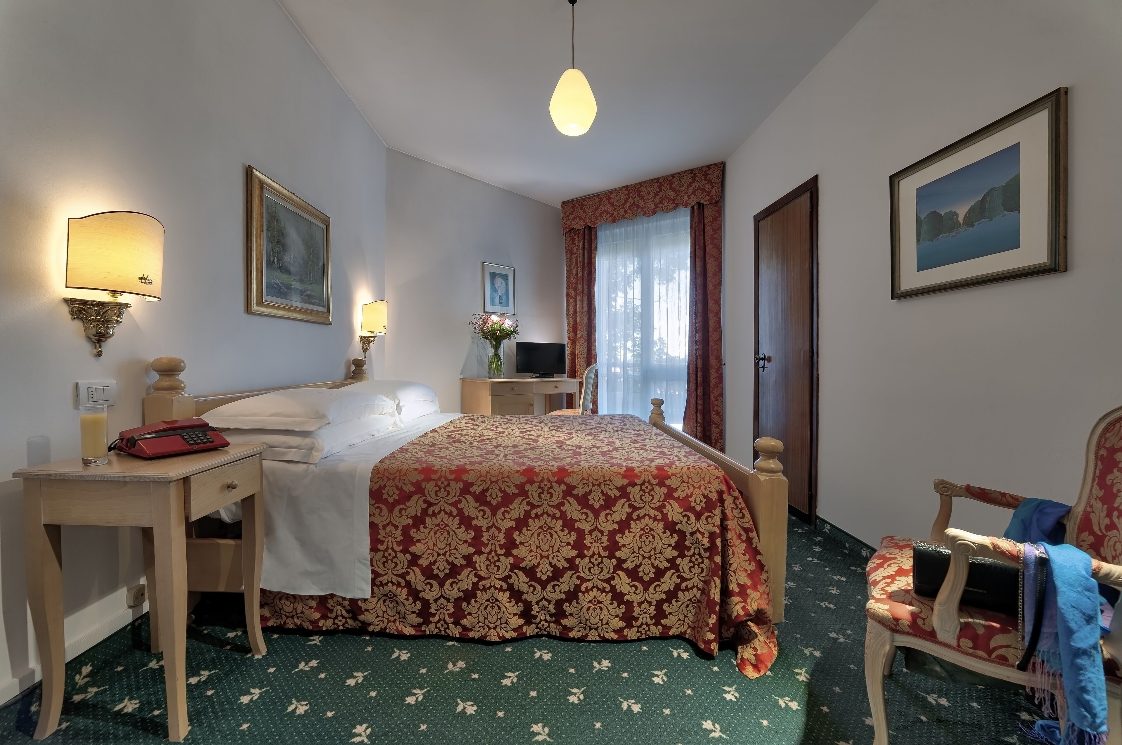 Una camera d'hotel con un letto, comodini, una scrivania, una sedia, una televisione e una finestra con tende.