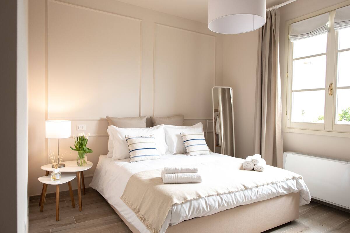 Een keurig opgemaakt bed met witte lakens en kussens, een nachtkastje met een lamp en een bloemenvaas, en een raam met gordijnen.