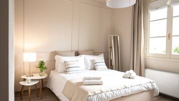 Una cama bien hecha con sábanas y almohadas blancas, una mesita de noche con lámpara y un jarrón de flores, y una ventana con cortinas.