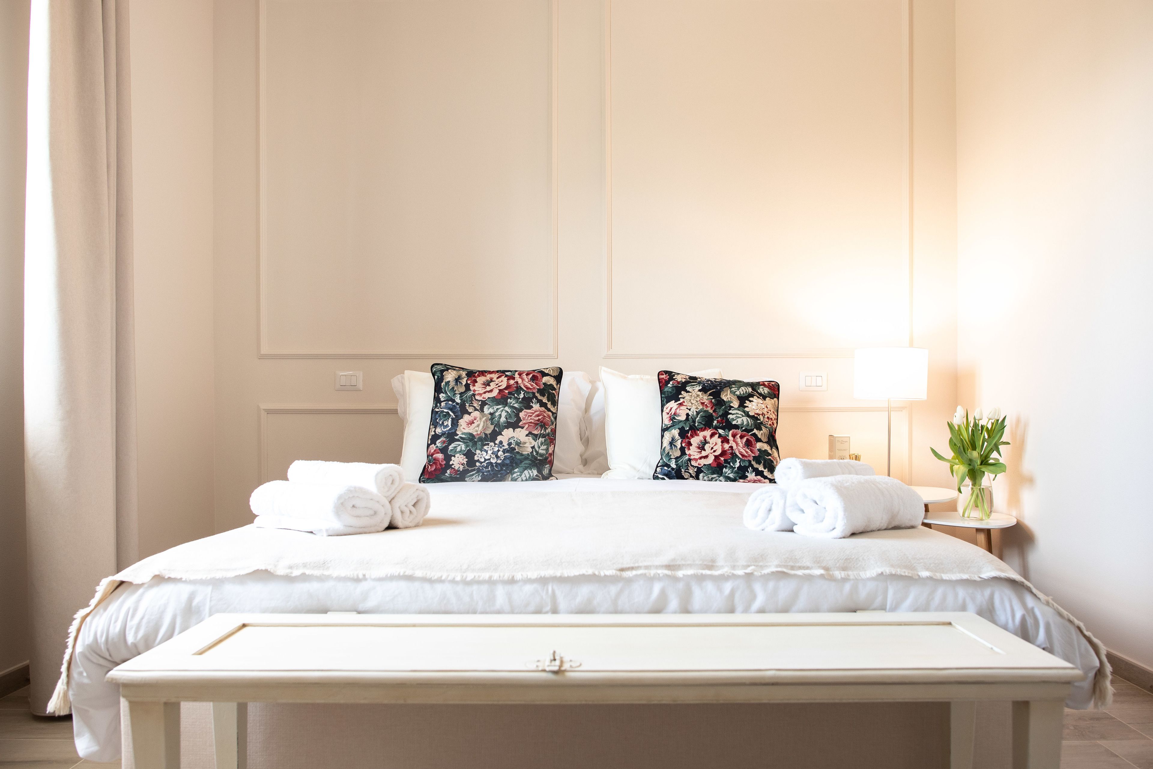 Une chambre d’hôtel avec un grand lit, deux coussins à motifs floraux, une table de chevet avec une lampe et un vase rempli de fleurs.