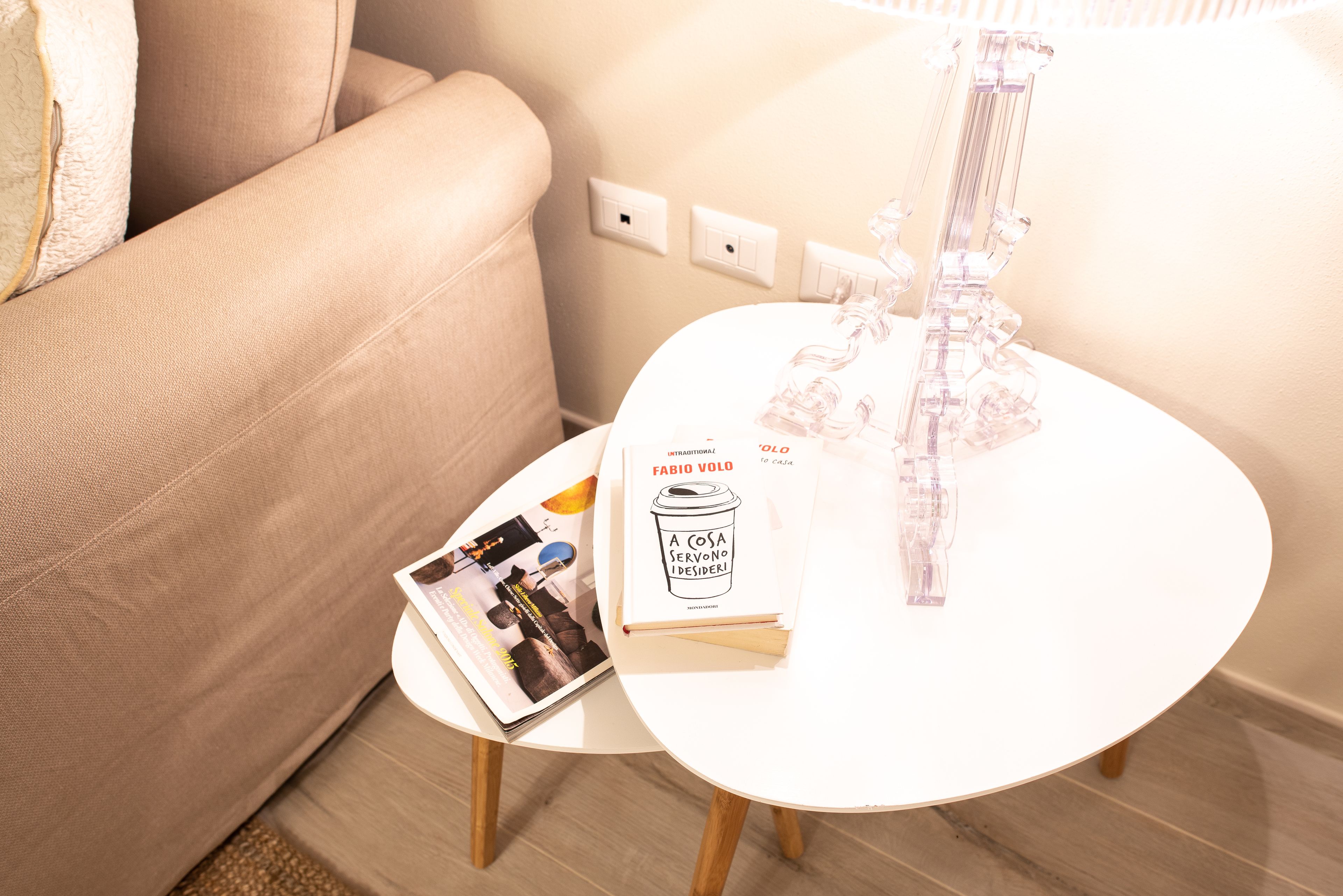 Una pequeña mesa blanca con un libro titulado 'A Cosa Servono I Desideri' y una revista, junto a un sofá color beige.