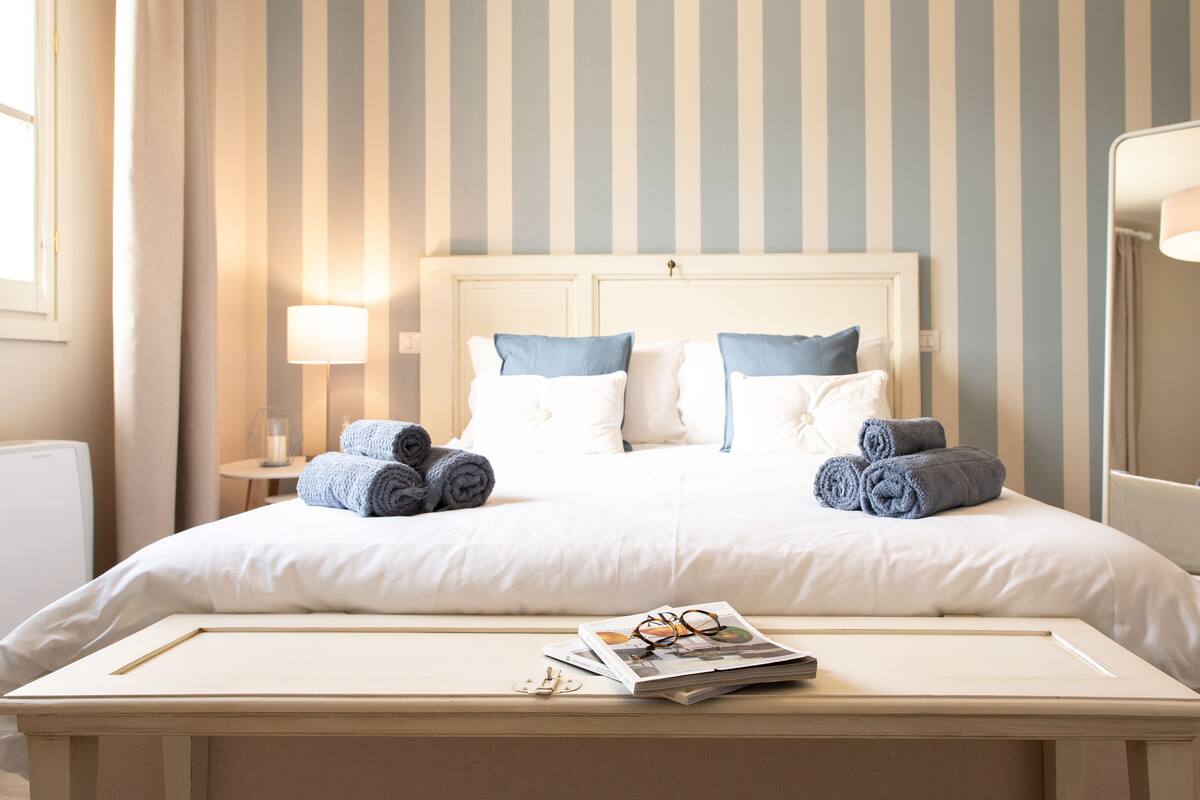 Een net opgemaakt bed met witte en blauwe lakens, een bedlampje en opgerolde handdoeken.