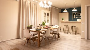 A dining area with a wooden table, white chairs, a chandelier, and a kitchen in the background. - La Locanda di Corrado (Cerreto Guidi)