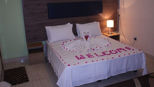 Una cama bien hecha, un tapete de bienvenida y una disposición de toallas con forma de corazón, una lámpara de noche y una mesita.